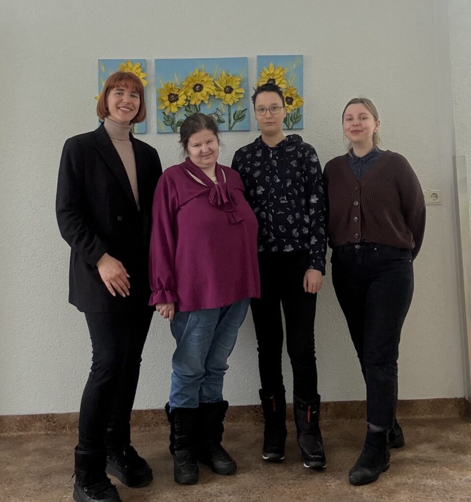 Prie sienos, ant kurios yra ryškių saulėgražų paveikslas, stovi 4 moterys: kairėje yra LSK regiono koordinatorė, Pagėgių SD rinkodaros specialistė Karolina Martišiūtė-El Oreibi, šalia jos dešinėje yra LSK vertintoja Jurgita Buivydaitė, LSK vertintoja Marija Amanovaitė, LSK koordinatorė Laura Mikelevičiūtė. Moterys pozuoja nuotraukai, šypsosi.