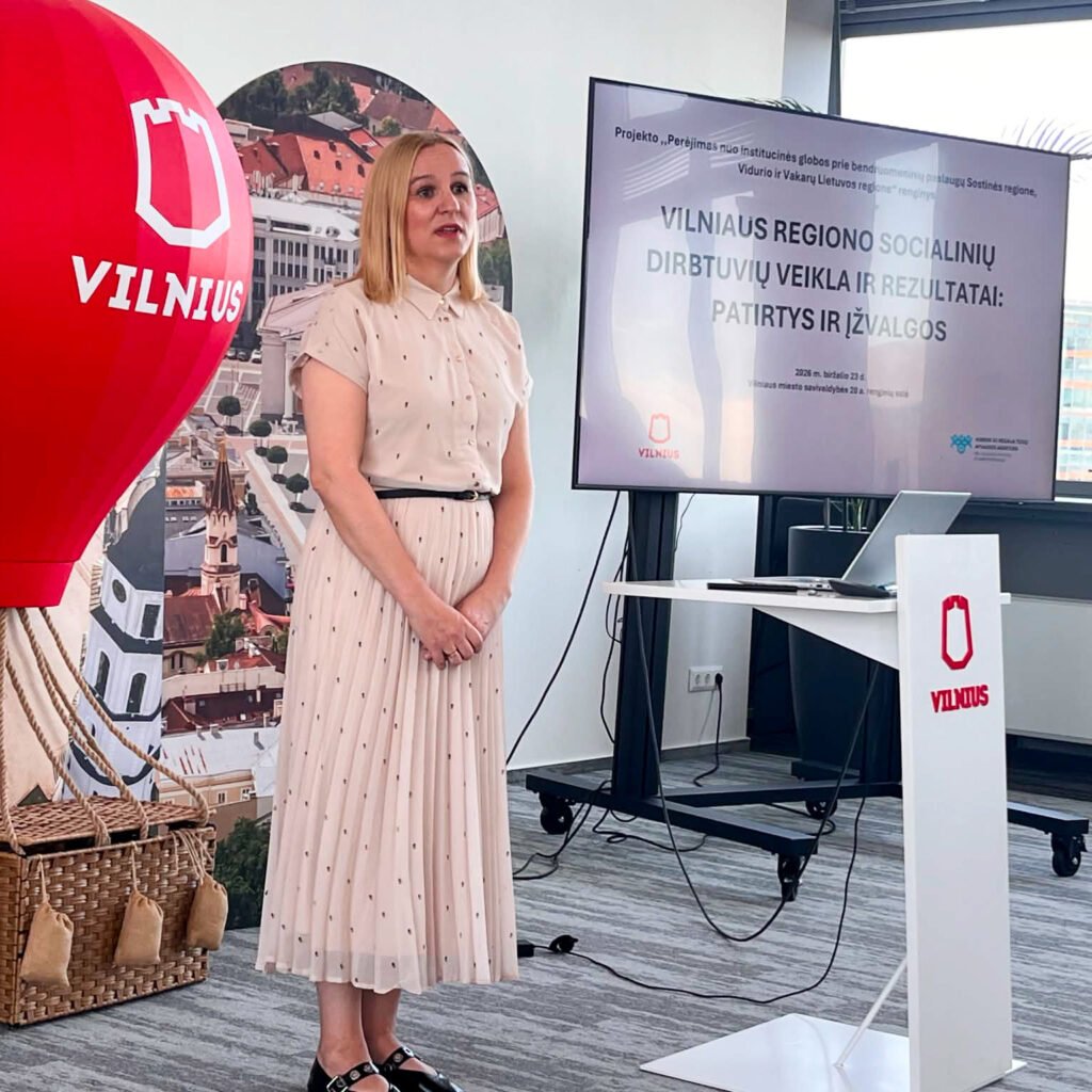 Šviesioje konferencijų salėje moteris stovi priekyje ir kalba renginio dalyviams. Ji vilki šviesią, kreminės spalvos suknelę su smulkiais taškeliais ir juodu dirželiu. Šalia jos stovi ekranas, kuriame rodoma prezentacija apie Vilniaus regiono socialinių dirbtuvių veiklą ir rezultatus. Kairėje matoma dekoratyvinė raudona oro baliono instaliacija su Vilniaus miesto logotipu, fone – miesto panoramos stendas. Dešinėje – tribūna su Vilniaus logotipu ir stalas su nešiojamuoju kompiuteriu. Salė moderni, gerai apšviesta, pro langus patenka dienos šviesa.