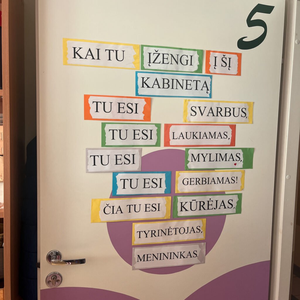Ant baltų durų pritvirtintas motyvuojantis užrašas iš spalvotų kortelių. Jame rašoma: „Kai tu įžengi į šį kabinetą, tu esi svarbus, tu esi laukiamas, tu esi mylimas, tu esi gerbiamas! Čia tu esi kūrėjas, tyrinėtojas, menininkas.“ Durų apačia dekoruota šviesiai violetiniais grafiniais elementais.