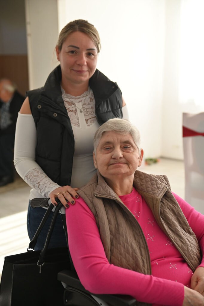 Šypsodamasi moteris stovi šalia vyresnės moters, sėdinčios neįgaliojo vežimėlyje, ir švelniai laiko ranką ant jos peties.